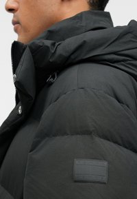 Juodas pūkinis džemperis su aukštu apykakle, užsegamas užtrauktuku ir spaudėmis. Textūruotas audinys ir guminis logotipo ženklelis ant rankovės.