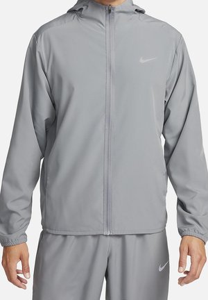 Homme portant une veste zippée Nike gris clair et un pantalon assorti avec des poignets élastiques et des logos Nike discrets sur la poitrine et la jambe.