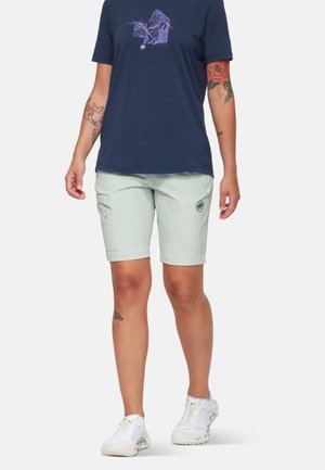 Person trägt ein marineblaues Grafik-T-Shirt, hellgraue knielange Shorts mit Seitentasche und weiße Sportschuhe, steht vor einem einfarbigen Hintergrund.
