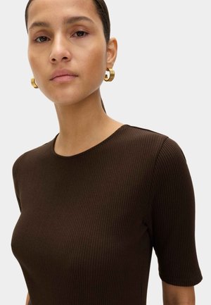 Donna che indossa una maglietta a maniche corte a coste di colore marrone scuro e orecchini a cerchio dorati, che guarda dritto con un'espressione neutra.