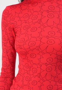 Rotes, geripptes Langarmoberteil mit einem gewagten schwarzen Blumenmuster, das abgerundete Blütenblätter und zentrale Kreise aufweist und ein strukturiertes Aussehen bietet.