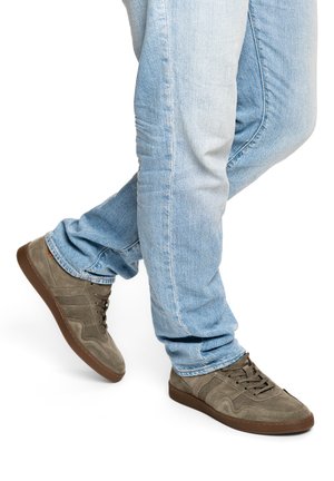 Hellblaue Jeans und braune Wildledersneaker mit Gummisohlen, ein Fuß leicht angehoben, vor weißem Hintergrund.