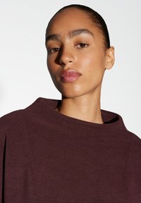 Bordeaux trui met een hoge, opgevouwen halslijn en geribbelde textuur. Gladde stof en subtiele ontwerpdetaillering benadrukken de minimalistische stijl.