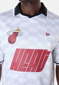 Witte sporttrui met zwarte kraag, voorzien van een rode "HEAT" grafiek en Miami-logo aan de voorkant, textuur met een gridpatroon op de achtergrond.