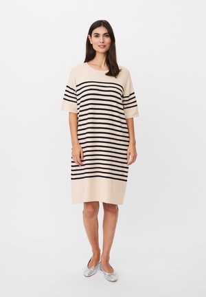 Donna in piedi che indossa un abito beige al ginocchio con strisce orizzontali nere e scarpe piatte argento su uno sfondo bianco semplice.