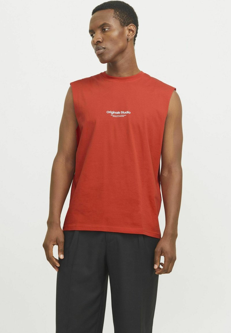 Rød ærmeløs t-shirt lavet af bomuld med rund halsudskæring, prydet med et hvidt trykt logo på forsiden, sammen med mørke bukser.