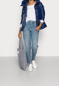 Person wearing blue puffer jacket, white shirt, light blue jeans, and white sneakers, holding a navy and white striped tote bag.