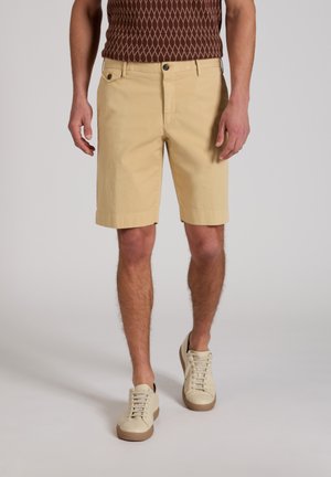 Mann trägt beige kniehohe Shorts, cremefarbene Sneakers und ein braunes, gemustertes ärmelloses Oberteil, steht vor einem schlichten Hintergrund.
