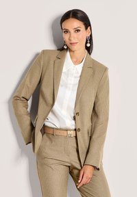 Beige gestructureerd pak met een getailleerde blazer en bijpassende broek. Gecombineerd met een witte blouse en een tan riem met een gouden gesp.