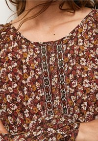 Blomsterbluse med brun bund og gule, røde og hvide blomster. Plisseret design og broderede detaljer langs udskæringen.
