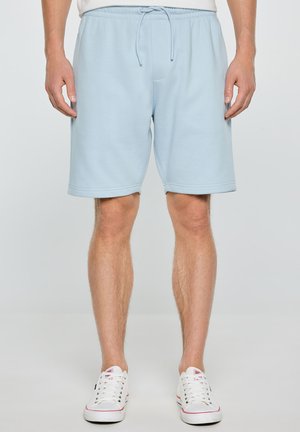 Vīrietis, kurš valkā gaiszilius šortus ar auklām un baltus sporta apavus, stāv pret vienkāršu fonu.