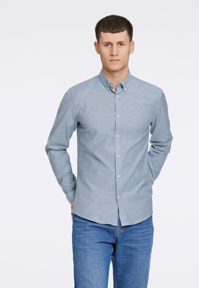 Camisa de botões azul clara em tecido de algodão, com colarinho clássico, mangas longas e barra reta, combinada com jeans azul de ganga.