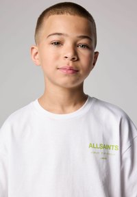 Balta kokvilnas T-krekls ar īsām piedurknēm, uz kreisās krūts ar zaļo logo, kas rakstīts "ALLSAINTS" kopā ar japāņu tekstu.