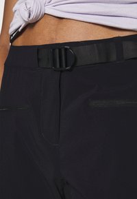 Gros plan d’un pantalon noir avec poches zippées et ceinture en tissu noir ajustable, porté sous une chemise blanche nouée révélant la taille.