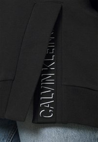 Felpa nera con spacco laterale e logo ricamato bianco "CALVIN KLEIN" su una patch nera. Tessuto misto cotone.