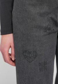 Σκούρες γκρι τζιν παντελόνες με μαύρα κεντημένα σχέδια από καρδιά, λουλούδι και αφηρημένα σχήματα, με κοντινή λήψη ενός χεριού πάνω στο ύφασμα.