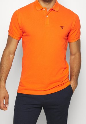 Mann trägt ein leuchtend oranges kurzärmeliges Polohemd mit kleinem Logo auf der Brust und dunkle Hosen, steht mit einer Hand in der Tasche.
