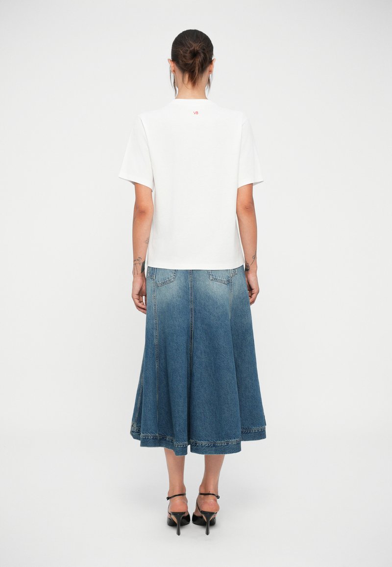 Vit t-shirt med kort ärm och en liten röd logotyp på ryggen, kombinerad med en lång ljusblå denimkjol. Modellen bär svarta strap-klackar.