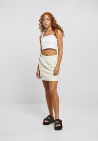 Top corto blanco combinado con una falda mini de cintura elástica en color crema y sandalias negras de suela gruesa. La modelo tiene el cabello rizado y está de pie contra un fondo neutro.