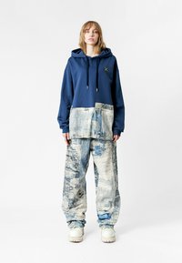 Sudadera azul marino con un bolsillo gráfico texturizado en azul y blanco; combinada con pantalones sueltos de un patrón azul claro y zapatillas blancas.