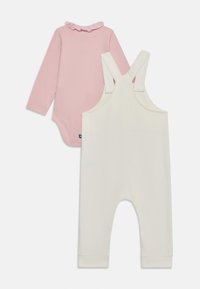 Rosa langærmet bodystocking med en flæsekrave, parret med hvide overalls med brede stropper og ribbede manchetter nederst.