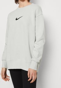 Ljusgrå sweatshirt med rund halsringning, ribbad vid ärmslut och nederkant. Har en svart Nike swoosh-logotyp på bröstet. Mjuk, avslappnad tyg.