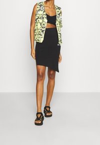 Svart asymmetrisk kjol kombinerad med en blommönstrad gul väst. Vesten har fickor och outfiten fulländas med svarta sandaler.