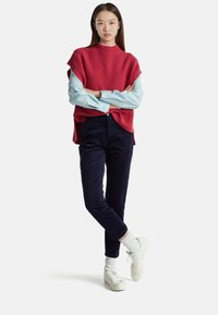 Red knit poncho over a light blue button-up shirt, paired with navy corduroy pants and white sneakers with ribbed socks. Casual outfit.
