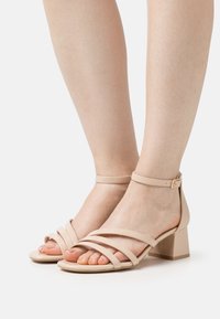 Beige hæle sandaler med remme, firkantet tå, lav blokhæl og ankelrem med detaljer i guld. Glat tekstur.