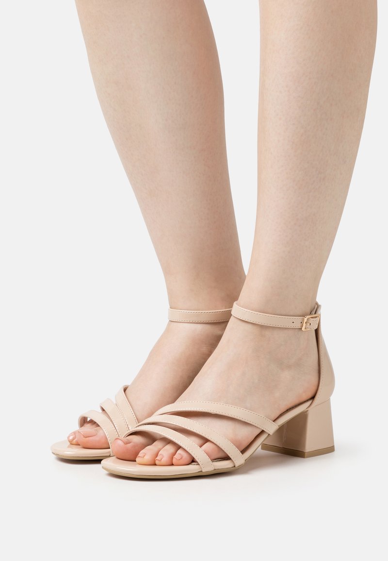 Beige hæle sandaler med remme, firkantet tå, lav blokhæl og ankelrem med detaljer i guld. Glat tekstur.