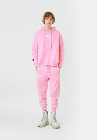 Sudadera rosa con cordones y un parche, combinada con pantalones de chándal a juego que presentan detalles bordados. Las botas de combate blancas completan el look.