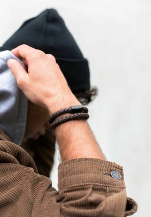 Rudas nerta odinė apyrankė su juodu metaliniu akcentu, dėvima ant riešo. Nešiojantis apsirengęs rudoje velveteen striukėje ir juodame kepure.