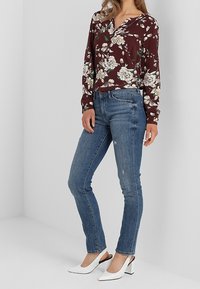 Blouse florale bordeaux avec un décolleté en V et des manches longues, associée à un jean skinny bleu. Des escarpins blancs à bout pointu rehaussent l'ensemble.