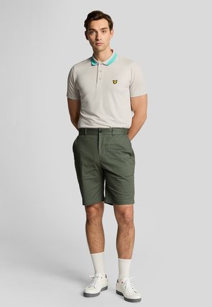 Lys grå polo shirt med turkis krave, med et gult logo. Oliven grønne shorts, hvide sneakers og hvide sokker.