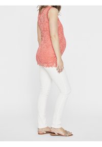 Korálová krajková blůza s vlnitým lemem, nošená přes bílé slim-fit kalhoty. Zobrazeno z boku na neutrálním pozadí. Sandály jsou béžové.
