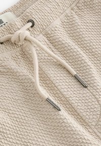 Beige teksturerede joggers med en tyk snor med metalspidser og et label i taljen. Overfladen har et ensartet vævet mønster.
