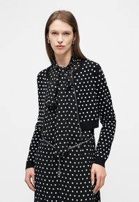 Γυναίκα που φοράει μαύρο διάφανο πουκάμισο με πουά και φιόγκο, κοντό πουλόβερ με μαύρα πουά και φερμουάρ, καθώς και ασημένια ζώνη αλυσίδα.