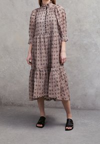 Vaalean taupe mekko, jossa on musta kukkakuosi, puffihihat, korkea kaulus, napit edessä ja kerroksellinen hame. Yhdistetty mustiin nauhakenkiin.