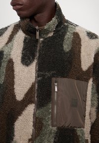 Giacca in pile con motivo camouflage marrone, verde e crema. Collo alto, zip frontale a metà e tasca sul petto con zip e dettaglio del marchio.
