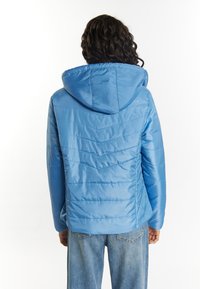 Blå huva-jacka med en kviltad design, som har en slät textur och snedställda sömmar. Modellen bär ljusblå jeans.