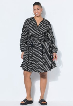 Frau steht und trägt ein längeres schwarzes Kleid mit Muster und Quasten an der Taille, schwarze Pantoletten und goldene Creolenohrringe, lächelnd.