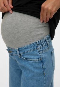 Pantalones de maternidad en denim azul claro con cintura elástica. Una blusa gris está metida por dentro, revelando una barriga redondeada.