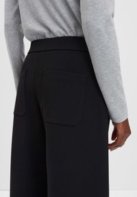 Zwarte high-waisted broek met twee rechthoekige achterzakken, gemaakt van een gestructureerde stof; gedragen met een lichtgrijze lange mouw top.