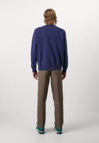 Felpa blu navy con polsini e orlo a costine, abbinata a pantaloni marrone chiaro con tasche laterali e scarpe da ginnastica teal.