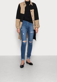 Leichtes, beiges und schwarzes farbblockiertes langes Hemd-Jackett über einem schwarzen Pullover, kombiniert mit stark ausgefransten blauen Skinny-Jeans und schwarzen Ballerinas.