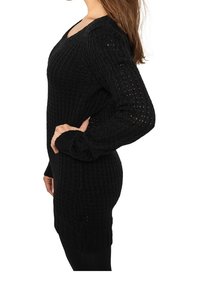 Femme portant une robe noire en maille à manches longues, se tenant de profil avec une main sur la hanche devant un fond blanc.