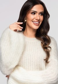 Femme aux longs cheveux ondulés foncés portant un pull blanc duveteux, souriante, touchant une boucle d'oreille en or sur un fond uni clair.