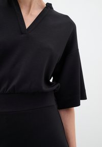 Crna majica s V-izrezom i kratkim rukavima, izrađena od mekog materijala. Ima opušteni kroj i sužen struk za elegantniji izgled.