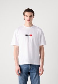 Balta kokvilnas T-kreklu ar īsām piedurknēm, uz kura ir vārds "HUG" melnā krāsā un sarkans sirds grafiskais attēls, veidots brīvā fitā.
