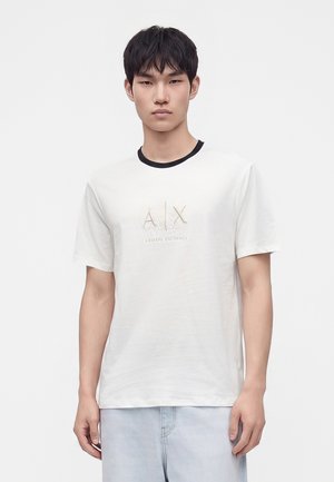 Ung mand iført en hvid Armani Exchange T-shirt med sort krave og lyseblå jeans, stående mod en enkel hvid baggrund.